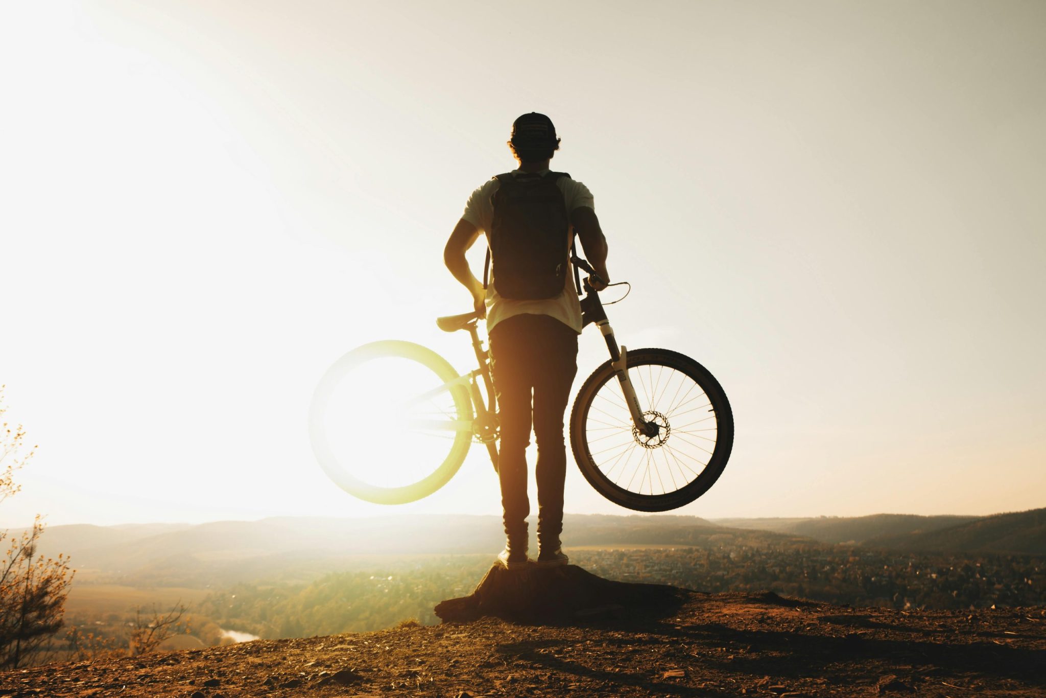 A cyclist stands silhouetted with a mountain bike on a hilltop during sunset, evoking adventure and freedom.